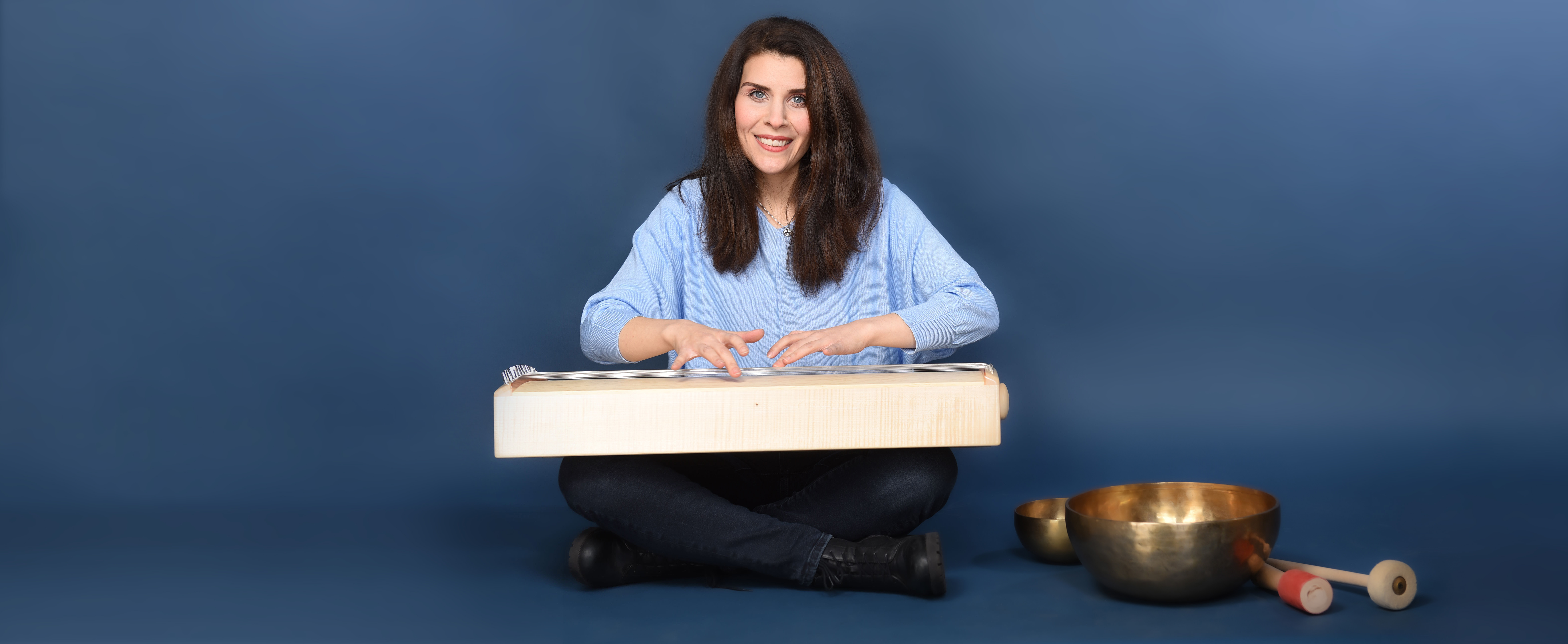 Fatma Koslik spielt ein Körpermonochord und lächelt. Neben ihr stehen Klangschalen – Musiktherapie für Entspannung und Wohlbefinden.